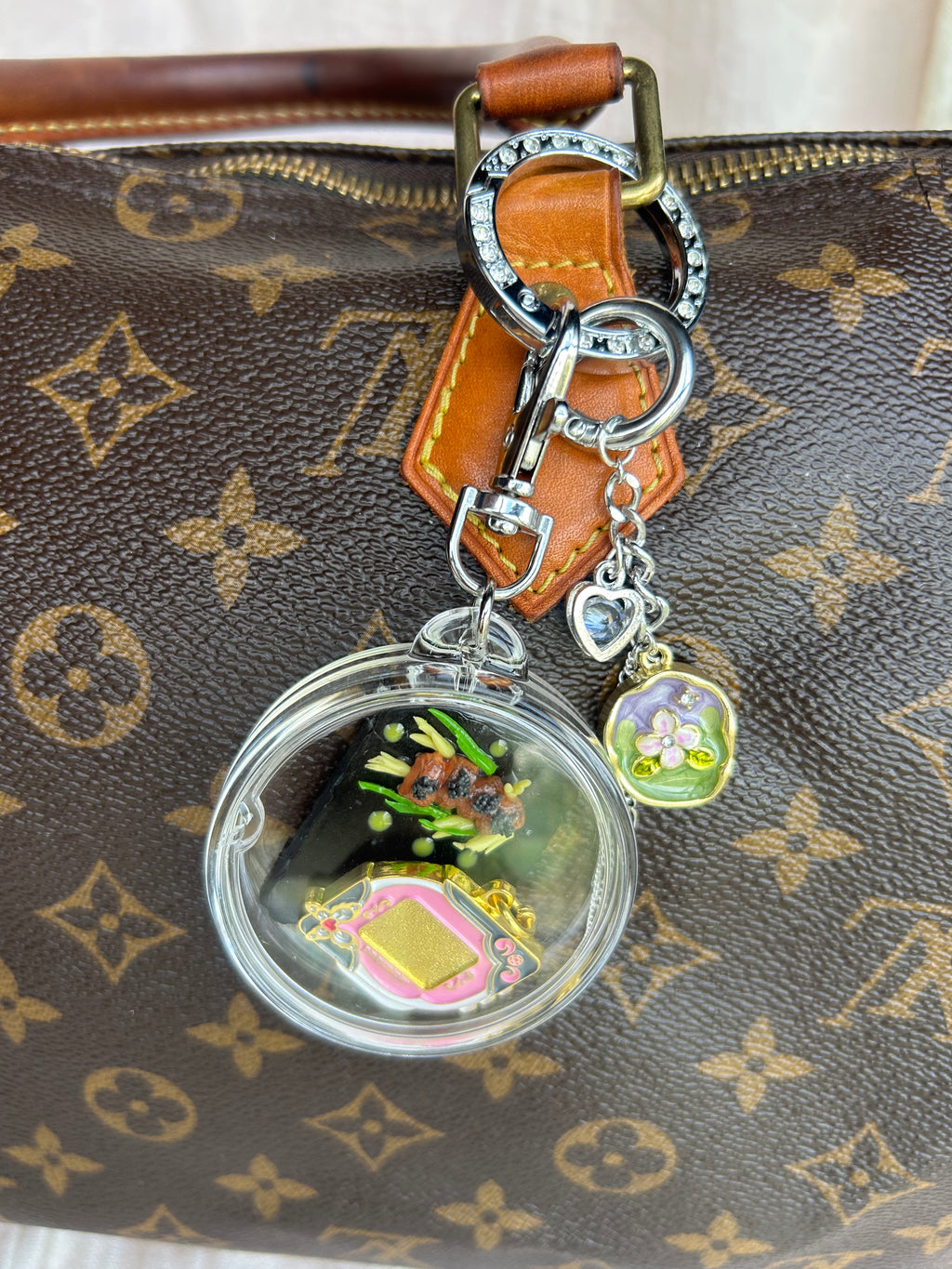 A Louis Vuitton monogram handbag in brown with gold-tone hardware, featuring a leather strap and a keychain-style charm with a round clear capsule containing colorful miniature objects and a small flower charm.