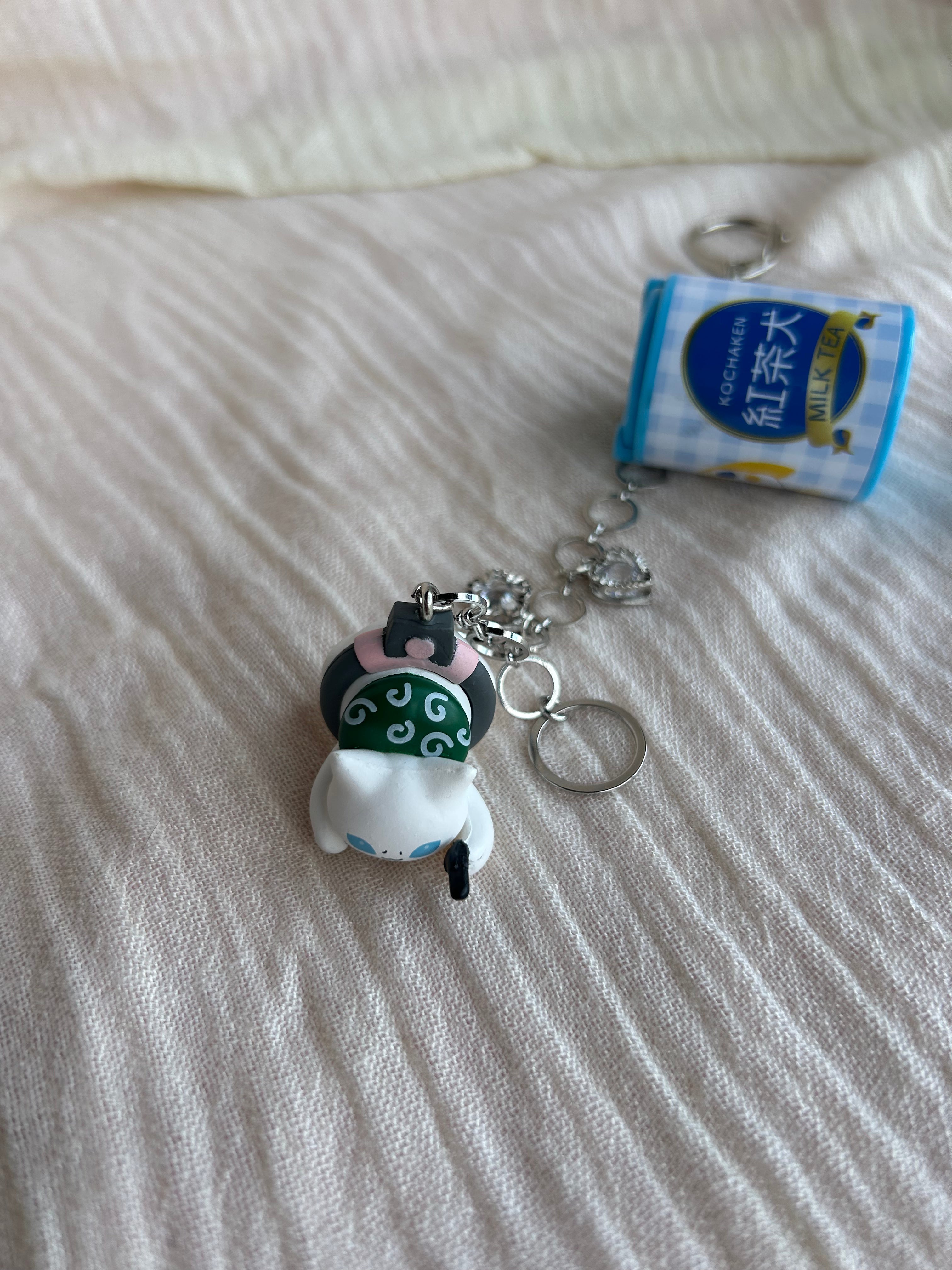 A small collectible keychain charm featuring a white cat wearing a green and black traditional mask, attached to a silver chain next to a miniature blue and white tea can keychain, resting on a textured off-white fabric surface.