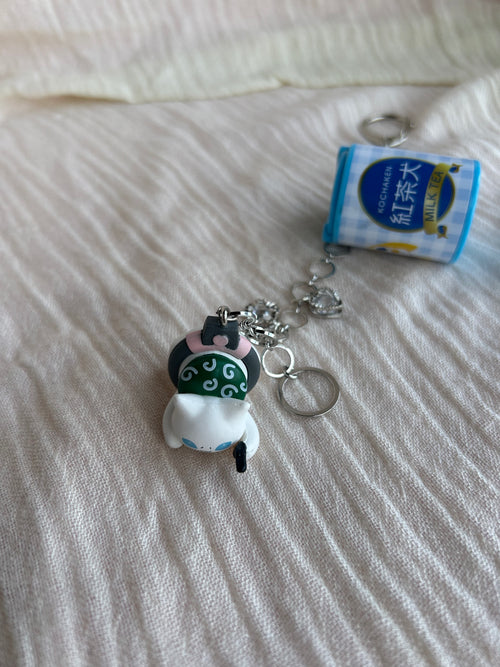 A small collectible keychain charm featuring a white cat wearing a green and black traditional mask, attached to a silver chain next to a miniature blue and white tea can keychain, resting on a textured off-white fabric surface.